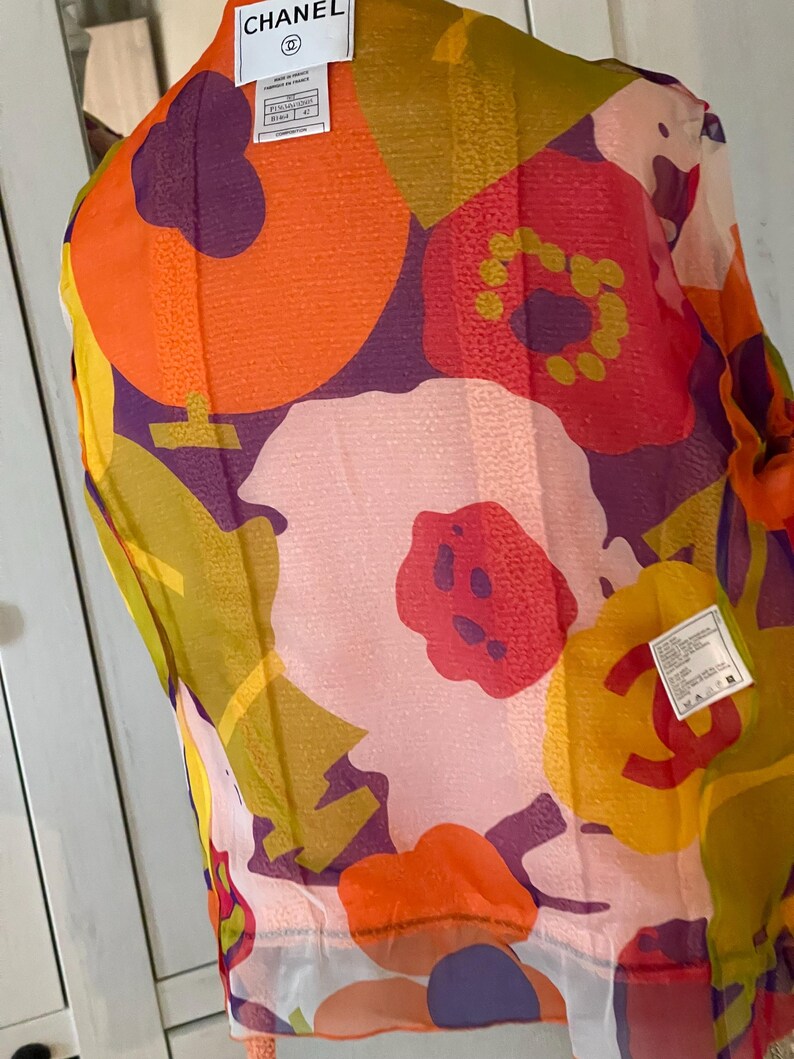 May include: A sheer scarf with a vibrant floral pattern in shades of orange, red, purple, yellow, and green. The scarf has a white border and a tag with the brand name "CHANEL".
