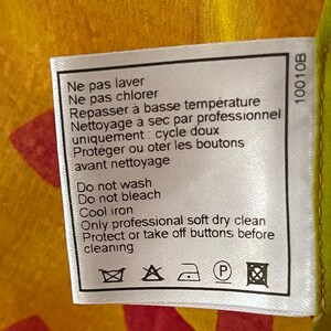 May include: A white fabric tag with care instructions in French and English. The tag includes symbols for washing, bleaching, ironing, and dry cleaning. The background is a mix of yellow, red, and green.