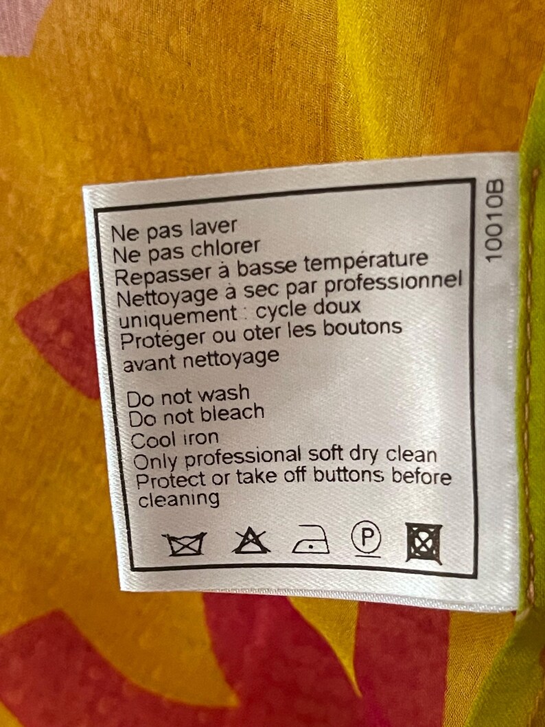 May include: A white fabric tag with care instructions in French and English. The tag includes symbols for washing, bleaching, ironing, and dry cleaning. The background is a mix of yellow, red, and green.