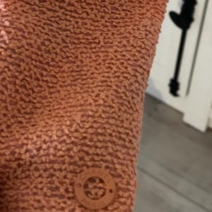 May include: Close-up of a textured, peach-colored fabric. A small, round, peach-colored emblem with the text "CHANEL PARIS" is visible. The fabric has a nubby, raised texture.