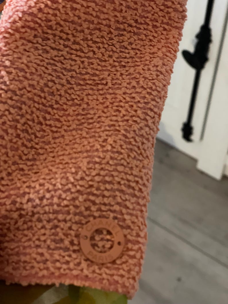 May include: Close-up of a textured, peach-colored fabric. A small, round, peach-colored emblem with the text "CHANEL PARIS" is visible. The fabric has a nubby, raised texture.