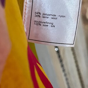 May include: A white fabric tag with black text indicating the material composition of a garment. The tag reads: 64% polyamide - nylon, 36% laine - wool, doublure/lining, 100% soie - silk. The tag is attached to a colorful fabric.