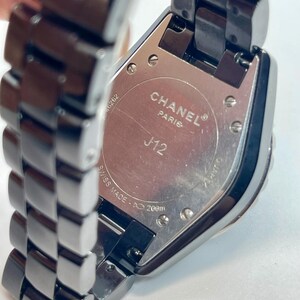 May include: A close-up of a black Chanel J12 watch. The watch features a rectangular face and a black metal strap. The back of the watch is visible, with the words "CHANEL PARIS J12 SWISS MADE" and "200m" printed on it.