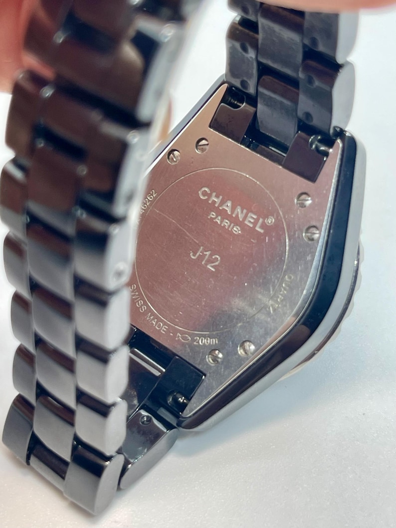 May include: A close-up of a black Chanel J12 watch. The watch features a rectangular face and a black metal strap. The back of the watch is visible, with the words "CHANEL PARIS J12 SWISS MADE" and "200m" printed on it.