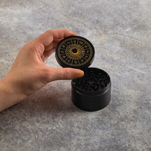 May include: Black grinder with a gold zodiac design on the lid. The lid is open and the grinder is on a gray surface.