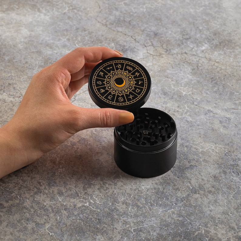 May include: Black grinder with a gold zodiac design on the lid. The lid is open and the grinder is on a gray surface.