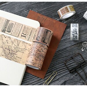 May include: Assortment of decorative washi tapes with vintage designs. The tapes feature a map, newspaper clippings, handwritten script, and numbers. The tapes are rolled up and displayed on a wooden surface.