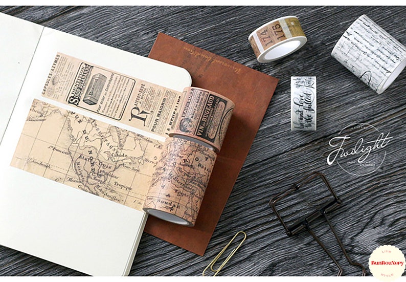May include: Assortment of decorative washi tapes with vintage designs. The tapes feature a map, newspaper clippings, handwritten script, and numbers. The tapes are rolled up and displayed on a wooden surface.