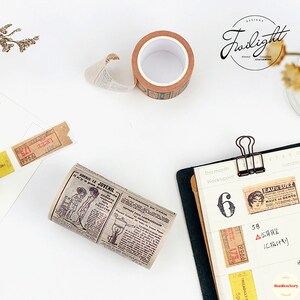 May include: A roll of brown washi tape with a vintage newspaper print design. The tape features a black and white illustration of a woman and text in French. The tape is on a white surface.