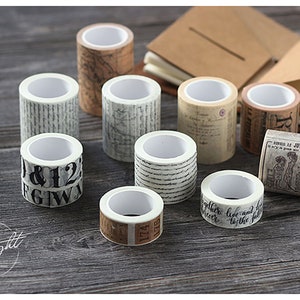 May include: Assortment of eight decorative washi tapes with various patterns, including numbers, script, and vintage illustrations. The tapes are rolled and displayed on a wooden surface.