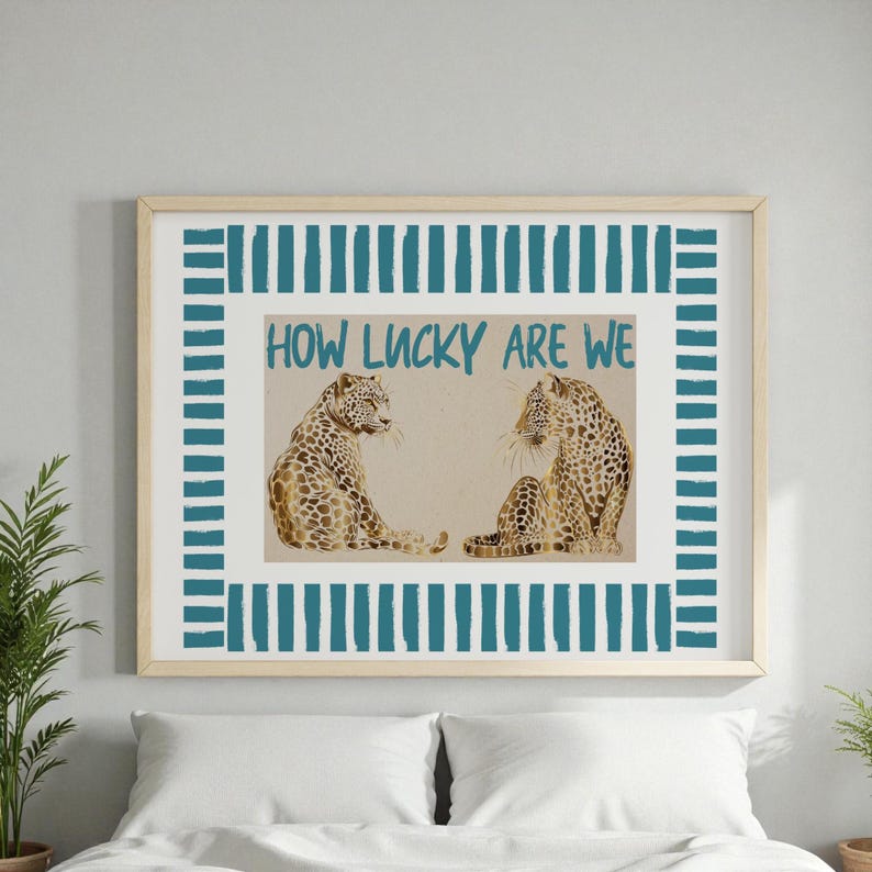 K&ouml;nnte beinhalten: Gerahmter Kunstdruck mit zwei Leoparden-Illustrationen mit braunen Flecken. Der t&uuml;rkisfarbene Text lautet "HOW LUCKY ARE WE", umgeben von einem t&uuml;rkis-wei&szlig; gestreiften Rand. Der Rahmen hat eine helle Holzfarbe.