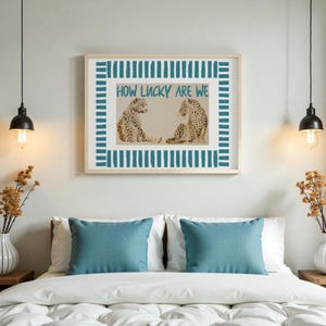 K&ouml;nnte beinhalten: Ein gerahmter Druck mit zwei Leoparden und dem Text "HOW LUCKY ARE WE" in T&uuml;rkis. Das Kunstwerk hat einen t&uuml;rkis-wei&szlig;en gestreiften Rahmen. Der Druck h&auml;ngt &uuml;ber einem Bett mit t&uuml;rkisfarbenen Kissen und einer wei&szlig;en Bettdecke.