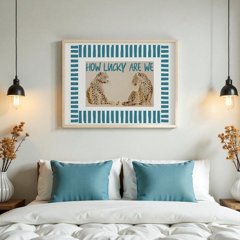 K&ouml;nnte beinhalten: Ein gerahmter Druck mit zwei Leoparden und dem Text "HOW LUCKY ARE WE" in T&uuml;rkis. Das Kunstwerk hat einen t&uuml;rkis-wei&szlig;en gestreiften Rahmen. Der Druck h&auml;ngt &uuml;ber einem Bett mit t&uuml;rkisfarbenen Kissen und einer wei&szlig;en Bettdecke.