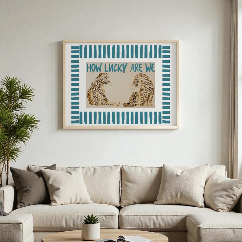 K&ouml;nnte beinhalten: Gerahmter Kunstdruck mit zwei Leoparden, die sich gegen&uuml;berstehen, mit dem Text "HOW LUCKY ARE WE" in T&uuml;rkis. Das Kunstwerk ist mit einem hellen Holzrahmen und einem t&uuml;rkisfarbenen Streifenrand gerahmt. Die Leoparden haben ein beige-braunes Fleckenmuster.