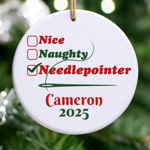 May include: White ceramic Christmas ornament with a brown string for hanging. The ornament features the words "Nice," "Naughty," and "Needlepointer" in red and green text. The name "Cameron" and the year "2025" are also displayed.
