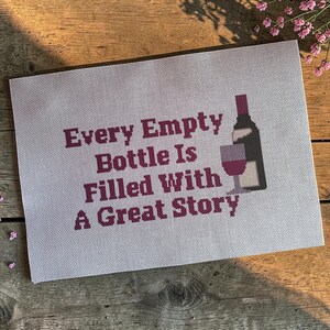 May include: A rectangular cross-stitch embroidery on white fabric featuring the dark red text Every Empty Bottle Is Filled With A Great Story, accompanied by a stitched graphic of a dark wine bottle and a glass filled with red wine.