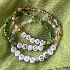 May include: Three beaded bracelets with green, white, and brown beads. The bracelets spell out the words "Faith", "Trust", and "Pixie Dust".