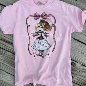 May include: Pink t-shirt with a graphic of Miss Piggy dressed in a pink dress and holding an umbrella. She is standing on pins and has a bow in her hair.