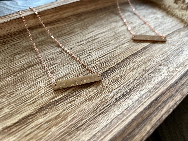 May include: Two gold necklaces with a bar pendant featuring a Mickey Mouse silhouette and stars. The necklaces are on a wooden surface.