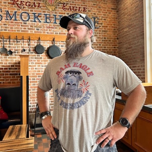 May include: A man wearing a gray t-shirt with a cartoon eagle design and the text "SAM EAGLE PRESIDENT". He is also wearing a black and white trucker hat and a black watch. The background features a brick wall and a sign that reads "REGAL EAGLE SMOKEHOUSE".