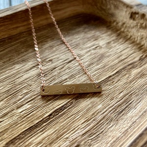 May include: A rose gold necklace with a bar pendant featuring three Mickey Mouse heads. The necklace is on a wooden surface.
