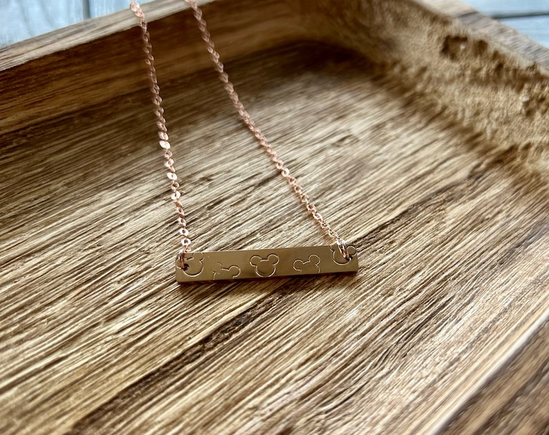 May include: A rose gold necklace with a bar pendant featuring three Mickey Mouse heads. The necklace is on a wooden surface.