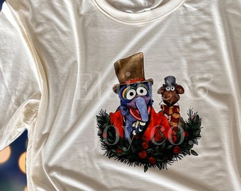 Può includere: T-shirt XL bianco sporco con una grafica colorata di due personaggi dei Muppet in abiti festivi. Il design include Gonzo con cappello a cilindro e cappotto rosso, e un cane marrone con cappello e sciarpa, circondato da una ghirlanda.