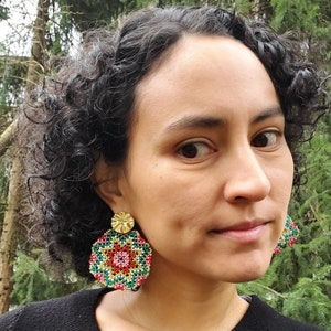 May include: A person wearing a black shirt with large, colourful, beaded earrings. The earrings are round and have a geometric pattern with red, green and gold accents.