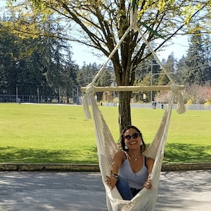 May include: A woman wearing sunglasses sits in a white hammock hanging from a tree branch. The hammock is made of a woven rope material. The woman is smiling and wearing a white tank top and blue jeans.