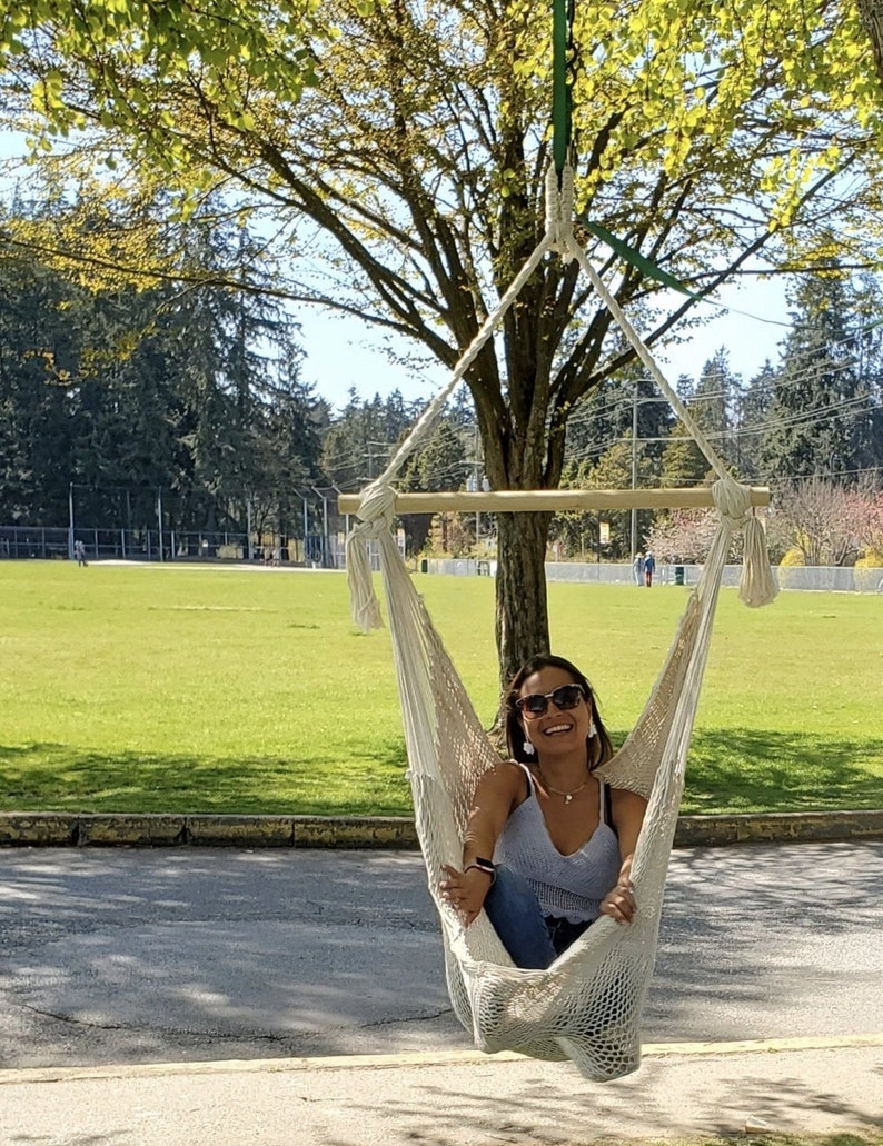 May include: A woman wearing sunglasses sits in a white hammock hanging from a tree branch. The hammock is made of a woven rope material. The woman is smiling and wearing a white tank top and blue jeans.