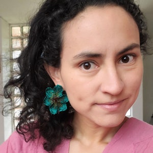 May include: A woman with curly dark hair wearing a pink shirt and turquoise flower-shaped earrings.