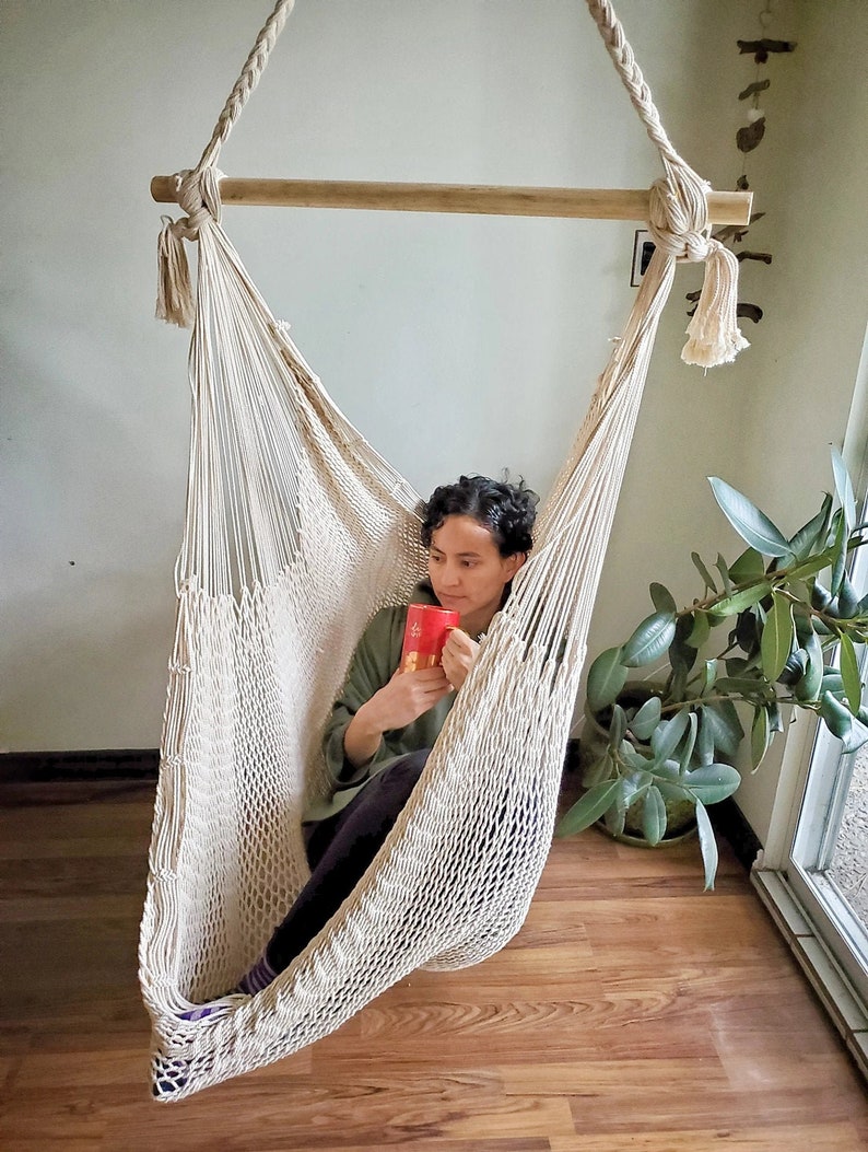 May include: A person relaxing in a white macrame hammock chair, holding a red mug. The hammock is hanging from a wooden bar and is suspended from the ceiling. The person is sitting in the hammock with their legs crossed and is looking off to the side.