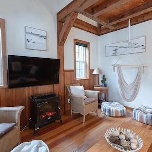 May include: A living room with a wooden floor, a fireplace, a television, a hammock, and two wicker chairs with cushions. The walls are white and the ceiling has exposed wooden beams.