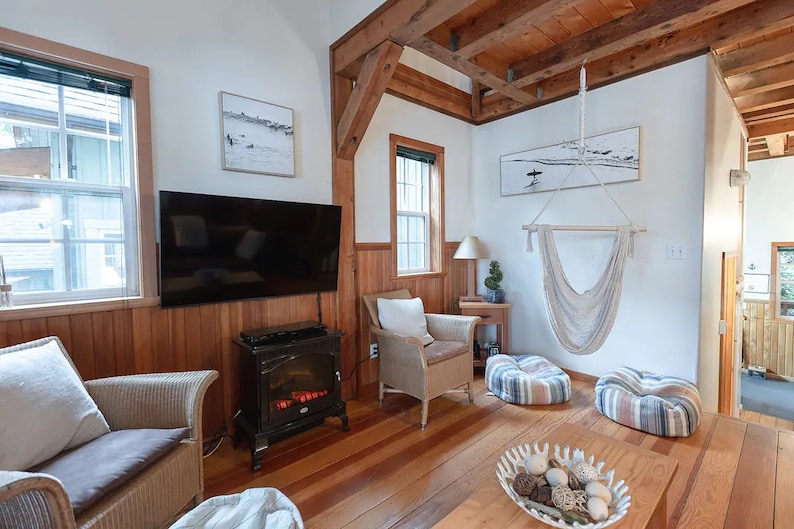 May include: A living room with a wooden floor, a fireplace, a television, a hammock, and two wicker chairs with cushions. The walls are white and the ceiling has exposed wooden beams.