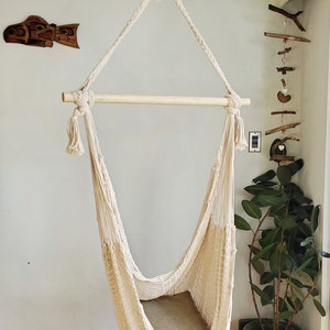 May include: A white cotton hammock chair with a wooden bar and tassels hangs from the ceiling. The hammock is empty and has a light brown cushion inside.