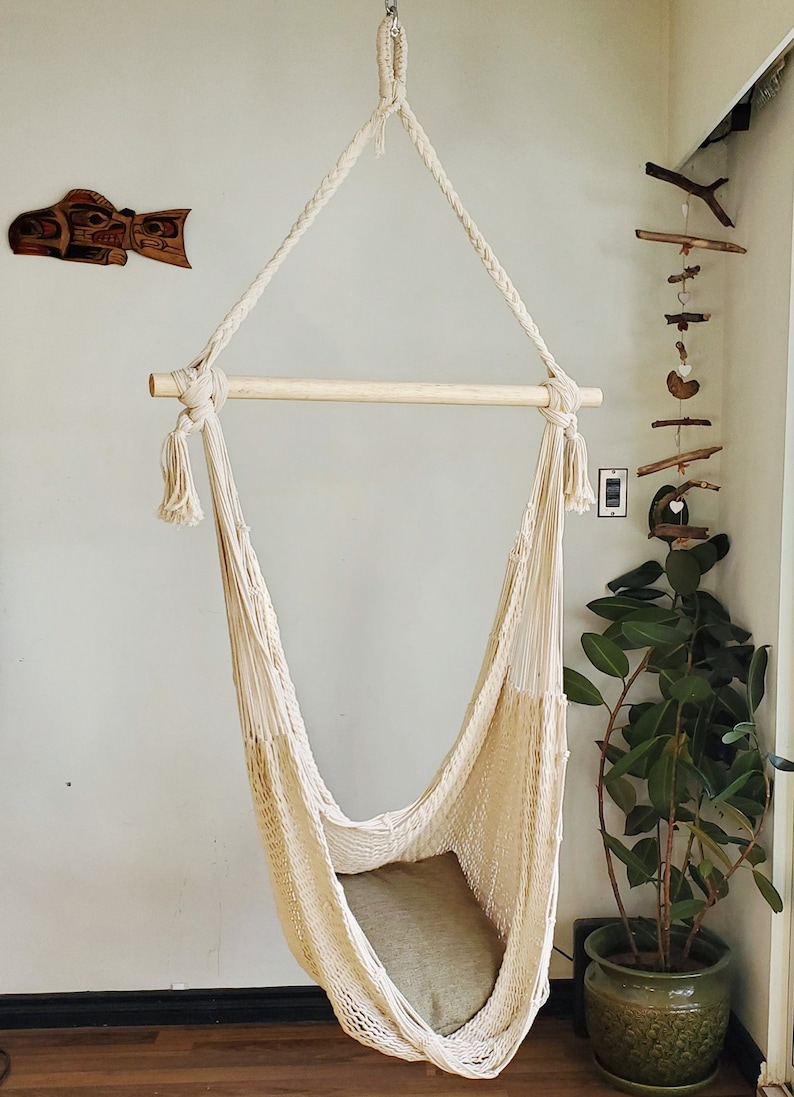 May include: A white cotton hammock chair with a wooden bar and tassels hangs from the ceiling. The hammock is empty and has a light brown cushion inside.
