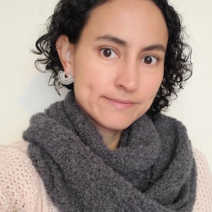 May include: A person wearing a grey, looped infinity scarf made of a textured, knitted material. The person is also wearing a light pink sweater and a silver earring. The scarf is wrapped around the neck.