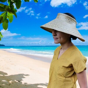 Op de afbeelding: Een geweven strooien vizier met een brede rand, met een patroon. Het vizier is bruin en beige, ontworpen om de ogen tegen de zon te beschermen. De achtergrond toont een strand met blauwe lucht en oceaan.