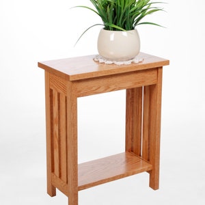 May include: A wooden side table with a natural finish. The table has a rectangular top, a lower shelf, and vertical supports. A white ceramic pot with green grass sits on top of the table. The table is approximately 24 inches tall.
