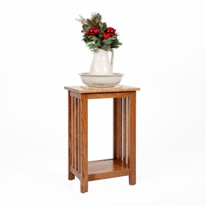 May include: A wooden plant stand with a single shelf. The stand is made of oak wood and has a natural finish. A white pitcher with a floral arrangement is on the top shelf.