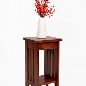 May include: A dark brown wooden side table with a square top and a lower shelf. A white vase with a ribbed texture holds a bouquet of orange berries. The table has vertical slats on the sides.