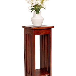 May include: A dark wood plant stand with a square top and a lower shelf. A white vase with a ribbed design holds a bouquet of white flowers. The stand is approximately 30 inches tall.
