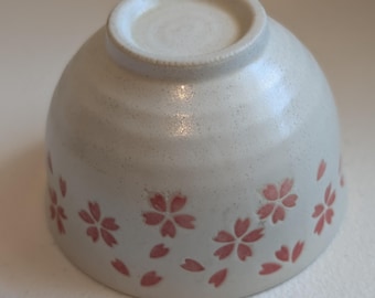 May include: A ceramic bowl with a speckled, off-white glaze. The bowl features a delicate design of pink cherry blossoms and falling petals around the exterior. The bowl is inverted, showing the base.