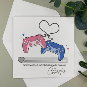 May include: A white card with a pink and blue video game controller design. The controllers are connected by a heart shape. The card reads "There's nobody I'd rather level up with than you" and is signed "Charlie".