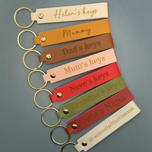 May include: A set of seven leather keyrings with gold key rings. Each keyring has a different colour and is personalised with a name and the word "keys" in gold lettering. The names on the keyrings are Helen, Mummy, Dad, Mum, Neve, Grandpa, and Ava.