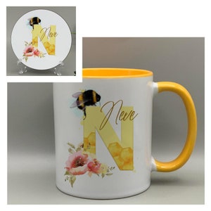 May include: A white ceramic mug with a yellow rim and handle. The mug features a watercolour illustration of a bee, flowers, and a honeycomb letter N. The text "Neve" is written in cursive on the mug.
