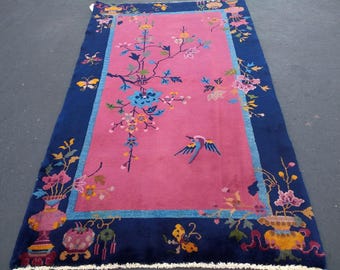 Può includere: Un tappeto rettangolare con un centro rosa e un bordo blu scuro. Il tappeto presenta disegni floreali e di uccelli in vari colori, tra cui blu, giallo e rosa. Il tappeto ha una frangia lungo il bordo inferiore.