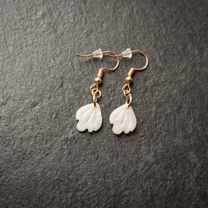 Könnte beinhalten: Ein Paar goldene Ohrringe mit weißen, blumenförmigen Anhängern. Die Ohrringe haben ein schlichtes Design und sind perfekt für den Alltag.