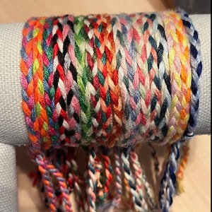 May include: A bundle of colorful friendship bracelets with various patterns and colors, including pink, blue, green, orange, and white. The bracelets are tied together at the top and have frayed ends.