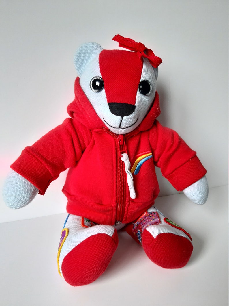 May include: A red and white plush teddy bear wearing a red hoodie with a rainbow zipper pull. The bear has black eyes and a black nose.